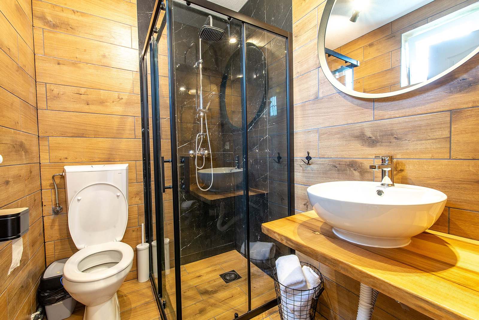 Kleine badkamer met houtlook wandtegels, douchecabine met zwart kader, toilet en een waskom op een houten blad
