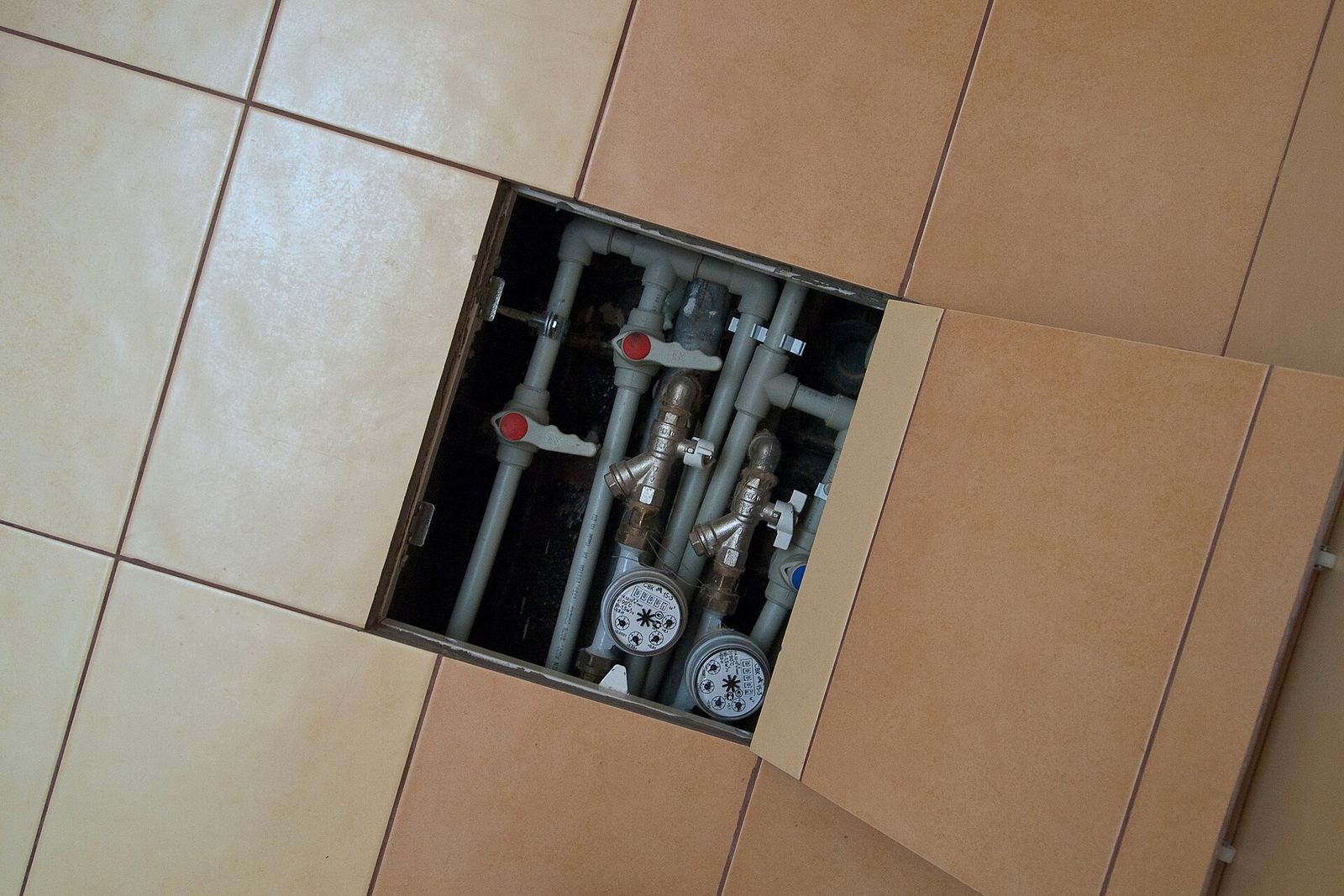 Opengeklapt tegelluik in een badkamerwand met kunststof waterleidingen, afsluitkranen en twee watermeters